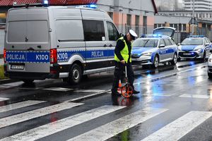 Na zdjęciu umundurowany policjant ruchu drogowego w żółtej odblaskowej kamizelce, za jezdni z urządzeniem służącym do mierzenia. W tyle policyjne radiowozy.