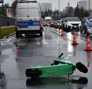 Na zdjęciu przewrócona na jezdni zielona hulajnoga, w tel policyjny radiowóz i inne pojazdy.