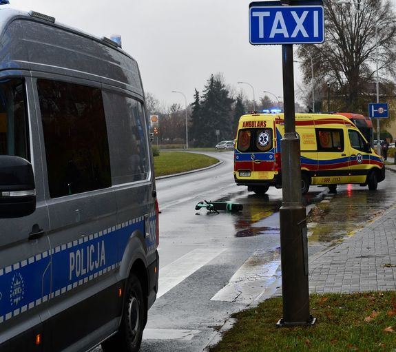 Na zdjęciu policyjny radiowóz, leżąca na jezdni hulajnoga, z nią ambulans pogotowia ratunkowego.