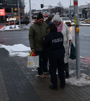 Na zdjęciu umundurowana policjanta rozdaje pieszym odblaskowe opaski.