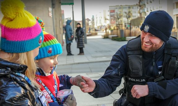 Na zdjęciu umundurowany policjant oraz dwoje dzieci ubranych w kolorowe czapki, jedno z nich trzyma w ręce puszkę Wielkiej Orkiestry Świątecznej Pomocy.