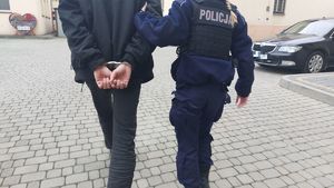 Na zdjeciu umundurowana policjantka lewą ręką prowadzi mężczyznę, który na ręce trzymane z tyłu ma założone kajdanki.