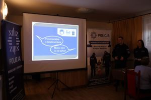 Na zdjęciu na środku tablica na której wyświetlony jest plakat debaty, z prawej i lewej strony roll up policji, z prawej stoi umundurowany policjant, obok niego umundurowana policjantka.