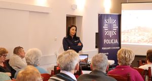 Na zdjęciu umundurowana policjantka stoi przed siedzącymi seniorami. Za nią rol up policji z napisem policja.