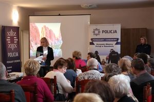 Na zdjeciu na środku sali stoi kobieta ubrana w czarny żakiet, za nią roll up policji z napisem policja oraz tablica z wyświetlonym pulpitem komputera. Na krzesełkach siedzą seniorzy.