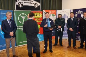Na zdjęciu w środkowej części p.o. Zastępca Komendanta Miejskiego Policji w Rzeszowie podinsp. Maciej Gawryś wręcza nagrodę chłopcu - jednemu z laureatów konkursu. Z prawej i lewej strony komendanta inni zaproszeni goście.