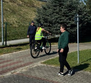 Na zdjęciu chłopiec w żółtej odblaskowej kamizelce jedzie na rowerze, z jego lewej strony stoi umundurowany policjant. Z prawej strony chłopiec w zielonej bluzie.