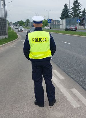 Na zdjęciu  stojący tyłem umundurowany policjant ruchu drogowego, w żółtej odblaskowej kamizelce z napisem policja na plecach.