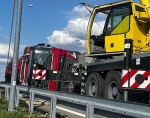 Na zdjęciu przewrócona na bok czerwona ciężarówka z wybita szyba kabiny, widoczne uszkodzenia blacharsko-lakiernicze. Przed nią stoi dźwig drogowy.