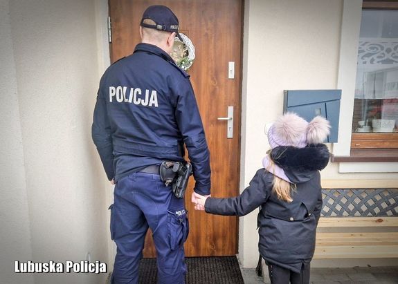 Na zdjęciu przed drzwiami wejściowymi do domu od strony zewnętrznej stoi umundurowany policjant, który za rękę trzyma dziewczynkę.