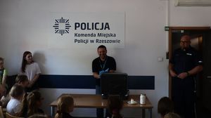 Na zdjeciu policjant technik kryminalistyki przeprowadza pokaz dla dzieci. Za min na ścianie tablica z napisem Komenda Miejska Policji w Rzeszowie.