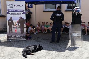 Na zdjeciu uczniowie SP nr 28 w Rzeszowie z wizytą w rzeszowskiej komendzie. Na środku odwrócony tyłem stoi policjant przewodnik psa, jego pie lezy na ziemi. Przed nimi siedzą dzieci.