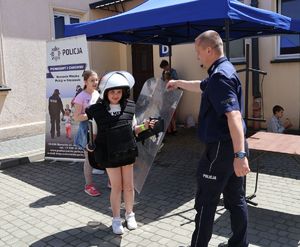 Na zdjeciu dziewczynka w policyjnej kamizelce  kamienioodpornej, na głowie ma założony kask, w ręce trzyma tarczę. Obok niej policjant.