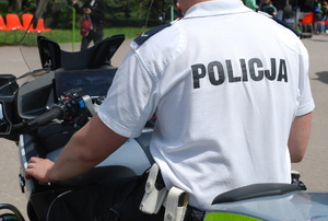 Na zdjęciu siedzący tyłem na motocyklu policjant w białej koszulce polo. Na plecach napis policja.
