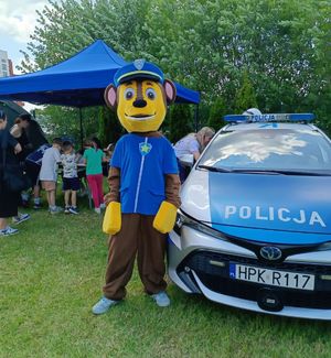 Na zdjęciu miś maskotka, obok policyjny radiowóz. w Tle stoisko profilaktyczne Komendy Miejskiej Policji w Rzeszowie.