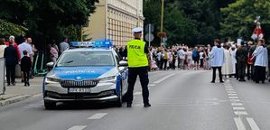 Na zdjęciu policyjny radiowóz, obok niego umundurowany policjant, w tle procesja.