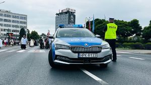 Na zdjęciu policyjny radiowóz, obok niego umundurowany policjant, w tle procesja.