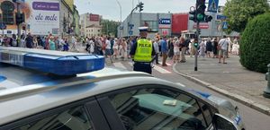 Na zdjęciu policyjny radiowóz,  przed nim umundurowany policjant, w tle procesja.