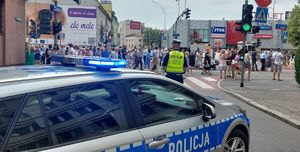 Na zdjęciu policyjny radiowóz,  przed nim umundurowany policjant, w tle procesja.