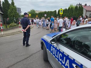 Na zdjęciu policyjny radiowóz,  przed nim umundurowany policjant, w tle procesja.