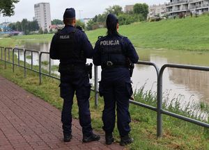 Na zdjęciu stoi odwróconych tyłem dwoje umundurowanych funkcjonariuszy policji, przed nimi rzeka.
