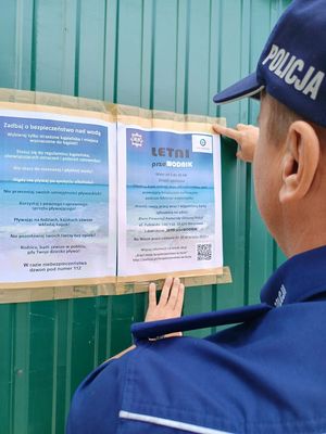 Na zdjęciu stojący tyłem policjant czyta zawieszone na ścianie budynku zasady bezpiecznego wypoczynku nad wodą.