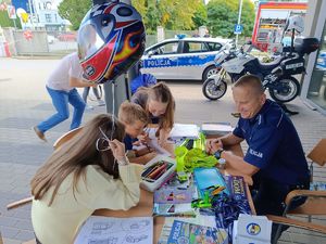Na zdjeciu umundurowany policjant siedzi przy stoisku profilaktycznym, obok niego dzieci.