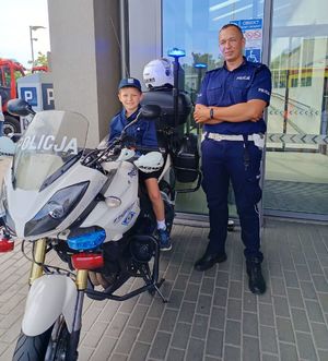 Na zdjeciu na policyjnym motocyklu siedzi chłopiec, na głowie na policyjny kask, obok niego stoi umundurowany policjant.