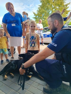 Na zdjęciu policjant przewodnik psa w umundurowaniu, obok niego leży policyjny pies. Przed nimi stoją dzieci i kobieta w niebieskim kitlu.