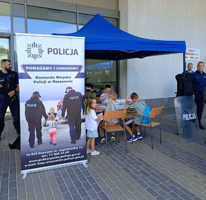 Na zdjeciu namiot z napisem policja, pod nim przy stolikach siedzą dzieck, obok stoi umundurowany policjant.