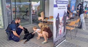 Na zdjeciu policja przewodnik psa trzyma psa na smyczy, którego dotyka trzymana na rękach kobiety mała dziewczynka.