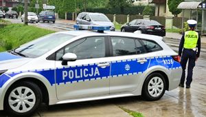 Na zdjeciu umundurowana policjanta ruchu drogowego, obok niej policyjny radiowóz.