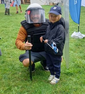 Na zdjeciu mężczyzna w policyjnym kasku trzyma tarczę. Obok niego stoi dziewczynka.