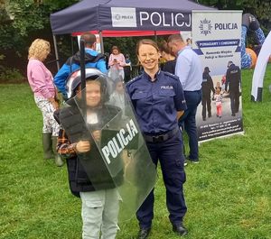 Na zdjęciu  umundurowana policjantka, obok niej chłopiec w policyjnym kasku, z tarcz a i pałką, z tyłu stoisko profilaktyczne Komendy Miejskiej Policji w Rzeszowie.