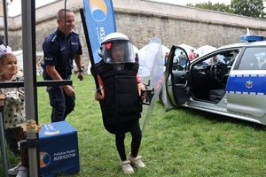 Na zdjęciu chłopiec ubrany w policyjny kask, kamizelę i tarczę, za nim umundurowany policjant, obok radiowóz.