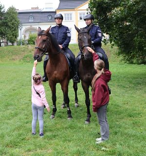 Na zdjęciu dwa policyjne konie służbowe, na których siedzą umundurowani policjanci. Dziewczynki głaskają konie.