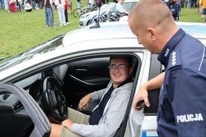 Na zdjęciu policyjny radiowóz, za kierownicą którego siedzi chłopiec i uśmiecha się obok stoi umundurowany policjant.