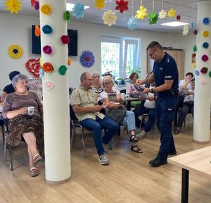 Na zdjęciu umundurowany policjant, na krzesełkach siedzą seniorzy.