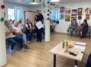 Na zdjęciu umundurowany policjant, na krzesełkach siedzą seniorzy.