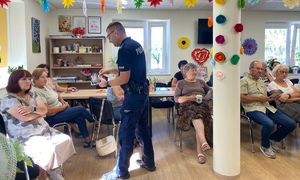 Na zdjęciu umundurowany policjant, na krzesełkach siedzą seniorzy.