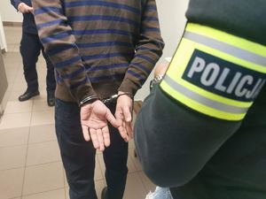 Na zdjęciu policjant w ubraniu cywilnym, na lewym przedramieniu złażoną ma opaskę z napisem policja. Przed nim, tyłem stoi mężczyzna, z założonymi na rękach kajdankami.