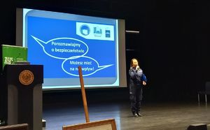 Na zdjęciu umundurowana policjantka z mikrofonem w ręce.Za nią na multimedialnej tablicy napis porozmawiajmy o bezpieczeństwie-możesz mieć n nie wpływ.