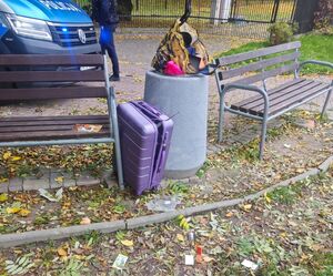 Na zdjęciu w betonowym koszu na śmieci walizka koloru fioletowego, za koszem policyjny radiowóz i umundurowany policjant.