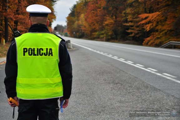 Na zdjęciu stojący tyłem umundurowany policjant, w żółtej odblaskowej kamizelce z napisem policja na plecach.