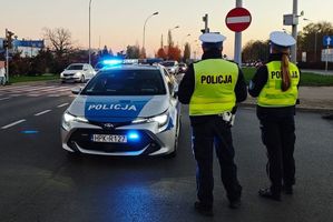 Na zdjęciu dwoje umundurowanych policjantów ruchu drogowego stających obok skrzyżowania. Przy nich policyjny radiowóz.