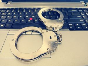 Na zdjęciu policyjne kajdanki leżą na klawiaturze laptopa.
