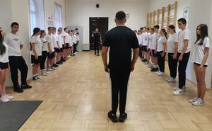 Na zdjęciu umundurowani policjanci w cywilnych ubraniach koloru czarnego. Przed nimi uczniowie klasy o profilu mundurowym, na zajęciach z elementów musztry.