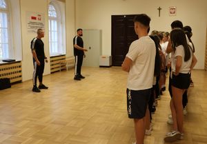 Na zdjęciu umundurowani policjanci w cywilnych ubraniach koloru czarnego. Przed nimi uczniowie klasy o profilu mundurowym, na zajęciach z elementów musztry.