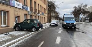 Na zdjęciu na pasie jezdni stoi policyjna furgonetka, po lewej stronie zielony pojazd przodem uderzył w elewację budynku.