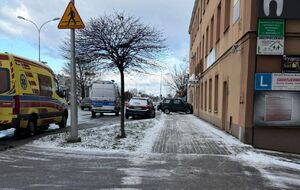 Na zdjęciu na pasie jezdni stoi policyjna furgonetka, po prawej stronie zielony pojazd przodem uderzył w elewację budynku.